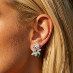 A close-up of a blonde woman wearing a pair of Emerald and Diamond Earclips with 1960s vintage design in 14K white gold, featuring round diamonds, marquise-cut stones in a floral shape, and a green emerald accent at the bottom.