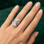 A hand with manicured nails wears the Diamond Ring, a platinum bow design set with old European diamonds, featuring vintage-inspired details against a dark green background.