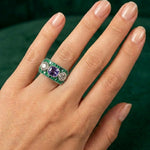 A hand with neatly manicured nails wears the Sapphire, Emerald and Diamond Ring: a wide 1930s platinum piece featuring a central emerald-cut purple sapphire, two round diamonds on each side, and a border of small green emeralds. The background is dark green fabric.