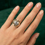A hand with neatly manicured nails wears a striking Rose Gold and Diamond Ring, featuring a prominent central gemstone, showcased against a dark green background.
