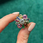 A hand with nude nails displays the Bezel Set Round Brilliant Diamond and Sapphire Multi Color Ring in Yellow Gold, featuring a central diamond surrounded by sapphires in various shapes and colors, set against a green background.
