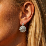Close-up of a person's ear with long blonde hair, wearing the 1930s Pair of Diamond Earrings featuring a sparkling, round cluster design set with old European-cut diamonds in platinum and gold that catch the light.