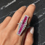 A hand showcases the Ruby and Diamond Ring—a large platinum piece with square-cut rubies bordered by small diamonds—set against a dark, textured background.
