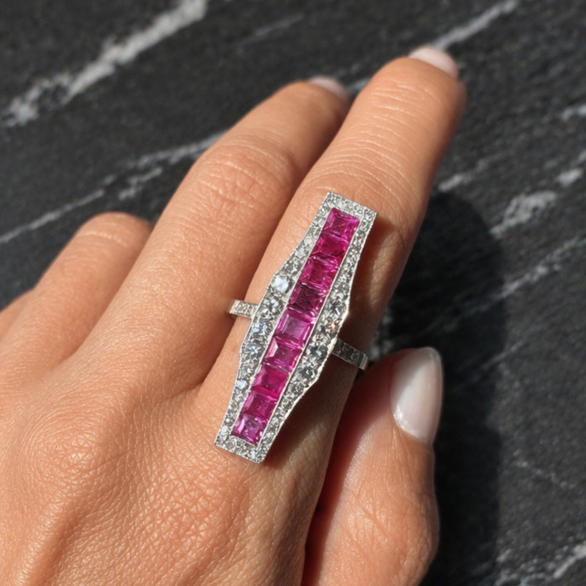 A hand showcases the Ruby and Diamond Ring—a large platinum piece with square-cut rubies bordered by small diamonds—set against a dark, textured background.
