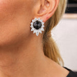 A close-up of a woman's ear wearing a Pair of Black Diamond and Diamond Earclips featuring large, flower-shaped platinum earrings with a black diamond center and sparkling white diamond accents.