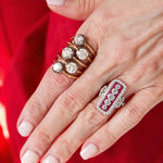 A hand with light pink nails wears multiple rings, including three gold rings with old European-cut diamonds on one finger and the Vintage Ruby and Diamond Ring—an ornate platinum and 18K white gold silver ring set with diamonds and calibré-cut rubies—on another, against a red fabric background.