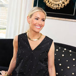 A smiling blonde woman in a studded black sleeveless top wears the Tiffany & Co. Tubogas Collar Necklace as she sits on a dark, studded sofa. Framed artwork and a curtain are visible in the background.