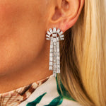Close-up of a woman's ear wearing a Pair of Diamond Earclips in platinum with baguette diamonds in a fan shape and three dangling rows. She has blonde hair and a patterned top, channeling the elegance of 1935.