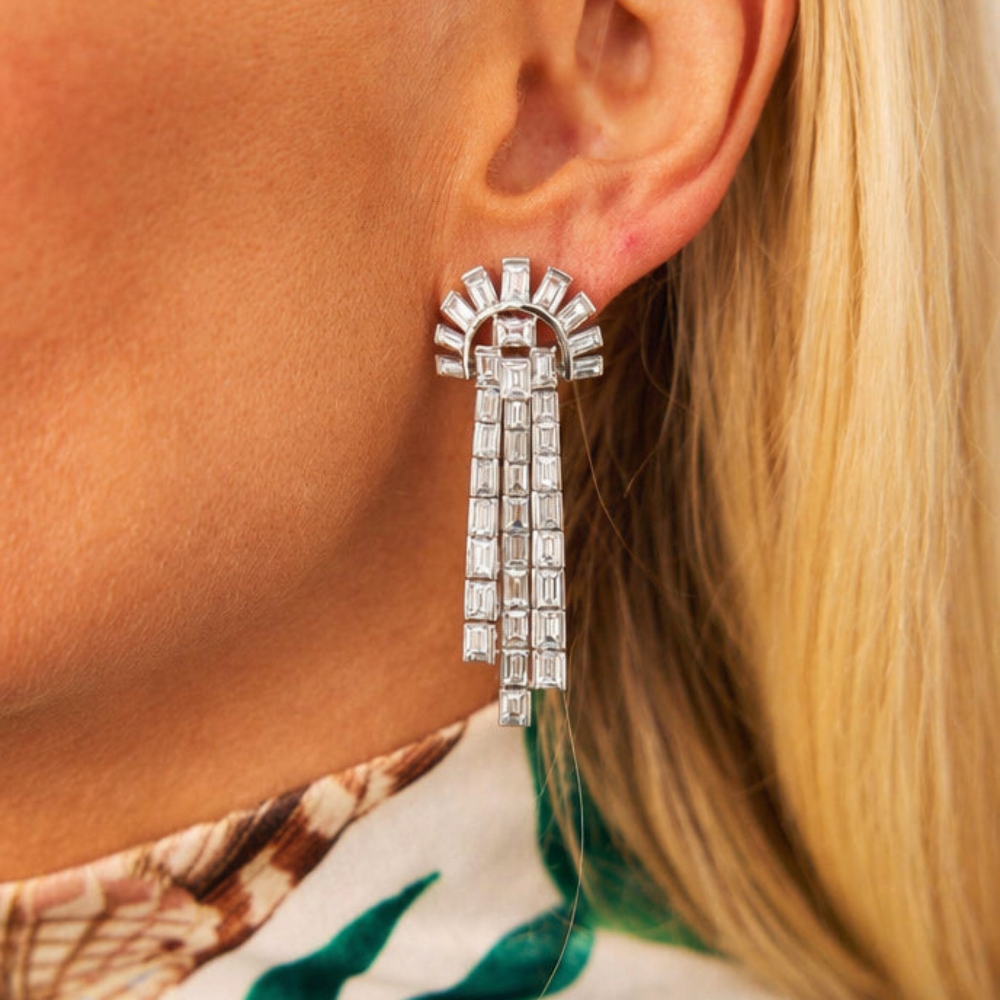 Close-up of a woman's ear wearing a Pair of Diamond Earclips in platinum with baguette diamonds in a fan shape and three dangling rows. She has blonde hair and a patterned top, channeling the elegance of 1935.
