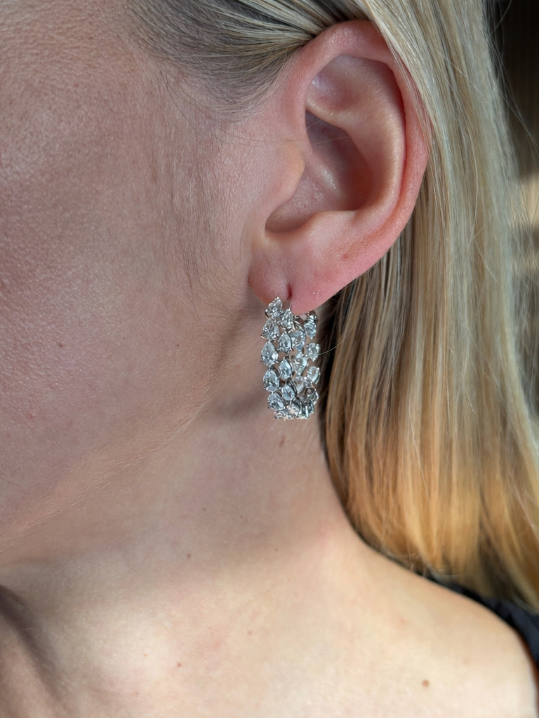 Close-up of a woman's ear wearing Duo Pear Shape Diamond Hoop Earrings in White Gold. Her blonde hair is tucked back, showcasing the sparkling elegance of these gemstone-adorned hoops.