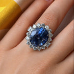 A hand adorned with the Vintage Sapphire and Diamond Ring, featuring a prominent oval Ceylon sapphire encircled by sparkling diamonds and set in radiant 18k white gold. The background is styled with softly draped yellow and beige fabric.