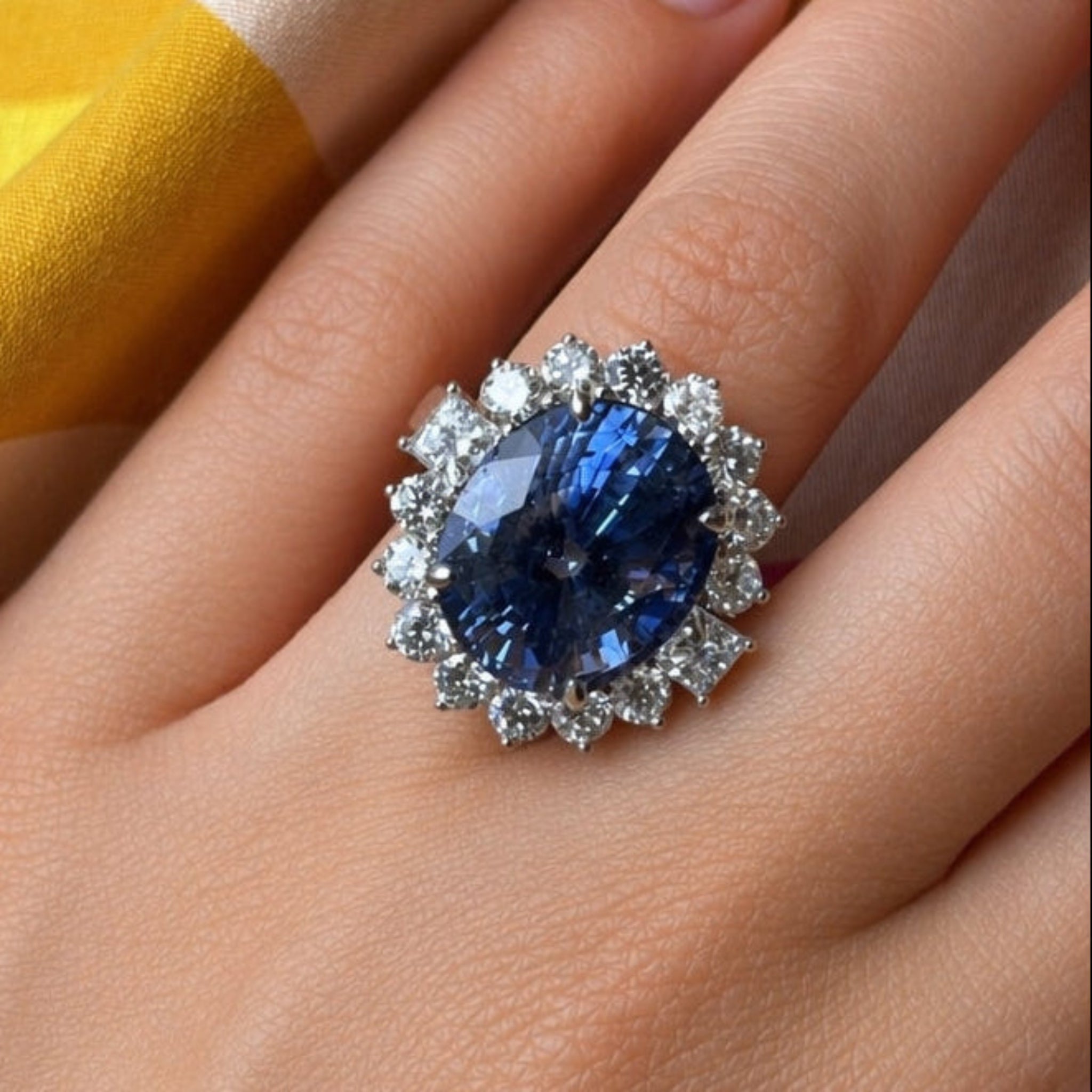 A hand displays the Sapphire and Diamond Ring, featuring a large oval Ceylon sapphire encircled by diamond accents in 18K white gold, set against a soft background with touches of yellow fabric.