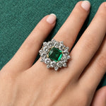 A hand with neatly manicured nails showcases the Vintage Emerald and Diamond Ring, featuring a square-cut Colombian emerald set in platinum and encircled by a halo of sparkling diamonds, displayed against a textured green background.