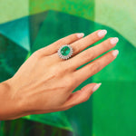 A hand with long, manicured nails wears the Vintage Emerald and Diamond Ring, showcasing a stunning central Colombian emerald encircled by sparkling diamonds and set in 18K white gold, all displayed against a vibrant green background.