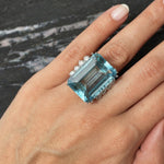 A Vintage Aquamarine and Diamond Ring, featuring a large rectangular emerald-cut aquamarine accented with round diamonds and set in platinum, adorns a person's finger. The hand rests on a dark marbled surface with light pink and white streaks, enhancing the ring’s brilliant setting.