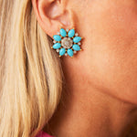 Close-up of a woman's ear wearing Marchak Turquoise and Diamond Earclips featuring large flower shapes with turquoise cabochons and gold petals. She has blonde hair and is wearing a pink and black top.