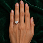 A hand with neatly manicured nails rests on dark green velvet, showcasing the Bezel Set Heart Shape Diamond Engagement Ring in Yellow Gold on the ring finger.