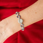 A woman's wrist showcases a Cartier Art Deco Sapphire and Diamond Watch with a round face, diamond accents, and blue gemstone links, set against a vibrant red background.