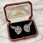 A pair of Cartier Diamond Earclips, circa 1950s, featuring a swirling leaf motif inspired by timeless Parisian elegance, rests in a red Cartier box lined with white satin and black velvet, displayed gracefully on a marble surface.