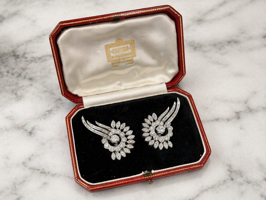 The Cartier Mid Century Diamond Earclips, featuring intricate swirling designs in platinum, are displayed in a red Cartier Paris jewelry box with black velvet and white satin lining on a marble surface.