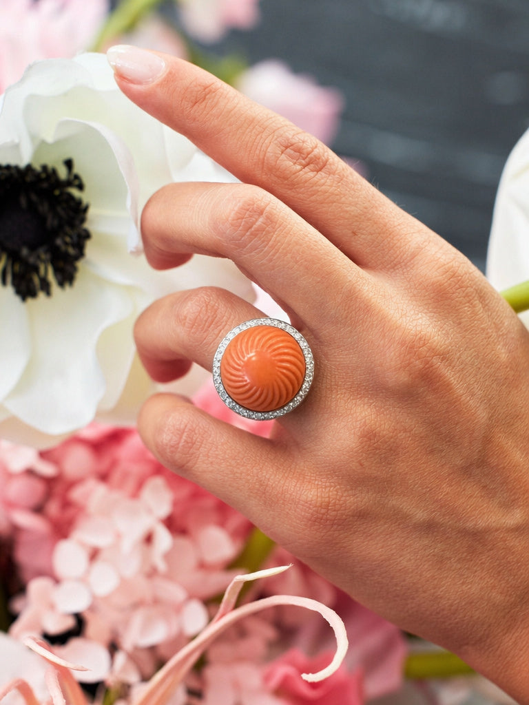 A hand wears the Cartier Carved Coral and Diamond Ring, featuring a large round orange coral with swirling patterns and pavé diamond border, set against white and pink flowers.