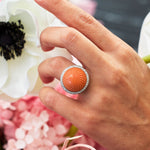 A hand wearing a Cartier Carved Coral and Diamond Ring, circa 1950s—featuring a large coral cabochon surrounded by pavé diamonds—is shown among white and pink flowers.
