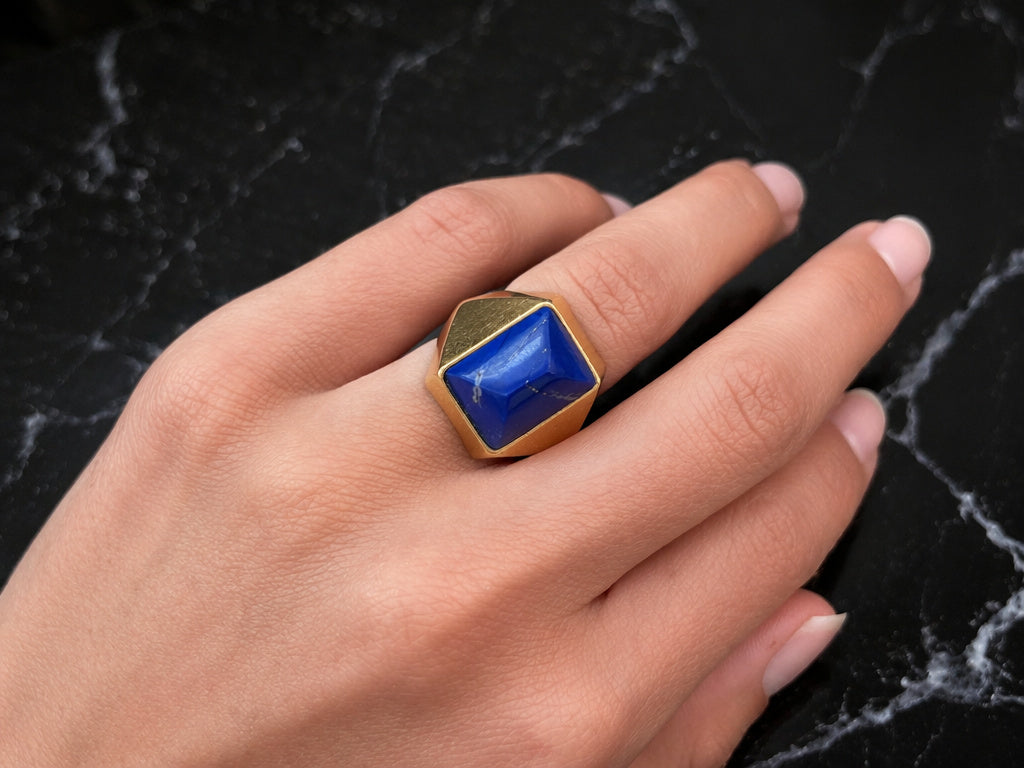 A hand with manicured nails wears the Boucheron Lapis Lazuli and Gold Ring, featuring a striking blue geometric stone set in 18k gold, elegantly displayed on a black marble surface.