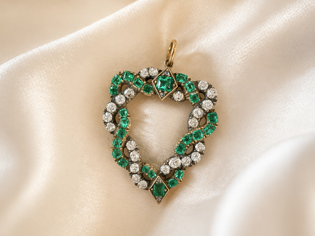 The Emerald and Diamond Pendant features cushion-shaped emeralds and old-cut diamonds set in gold, displayed on cream-colored satin fabric.