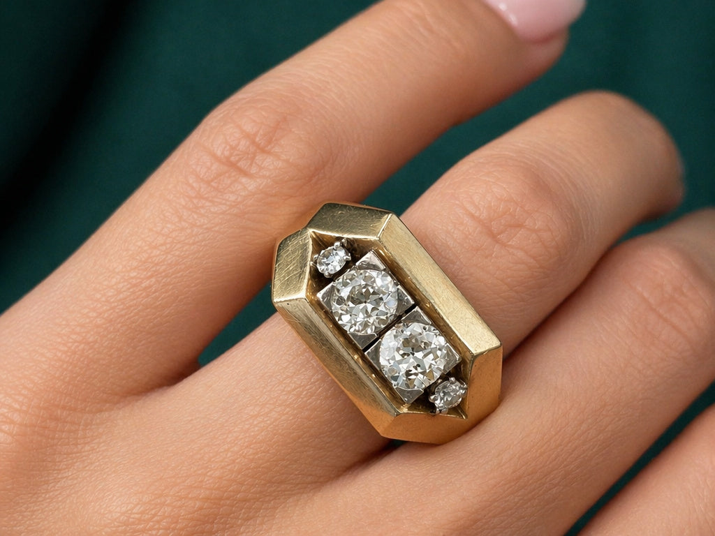 A close-up of a hand wearing the Mauboussin Gold and Diamond Ring with three large center diamonds and two smaller diamonds on each end, set against a dark green background, nails perfectly manicured.