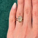 A hand with light skin wears the Double Band North-South Prong Set Cushion Cut Diamond Engagement Ring in yellow gold on the ring finger, shown against a textured green background.