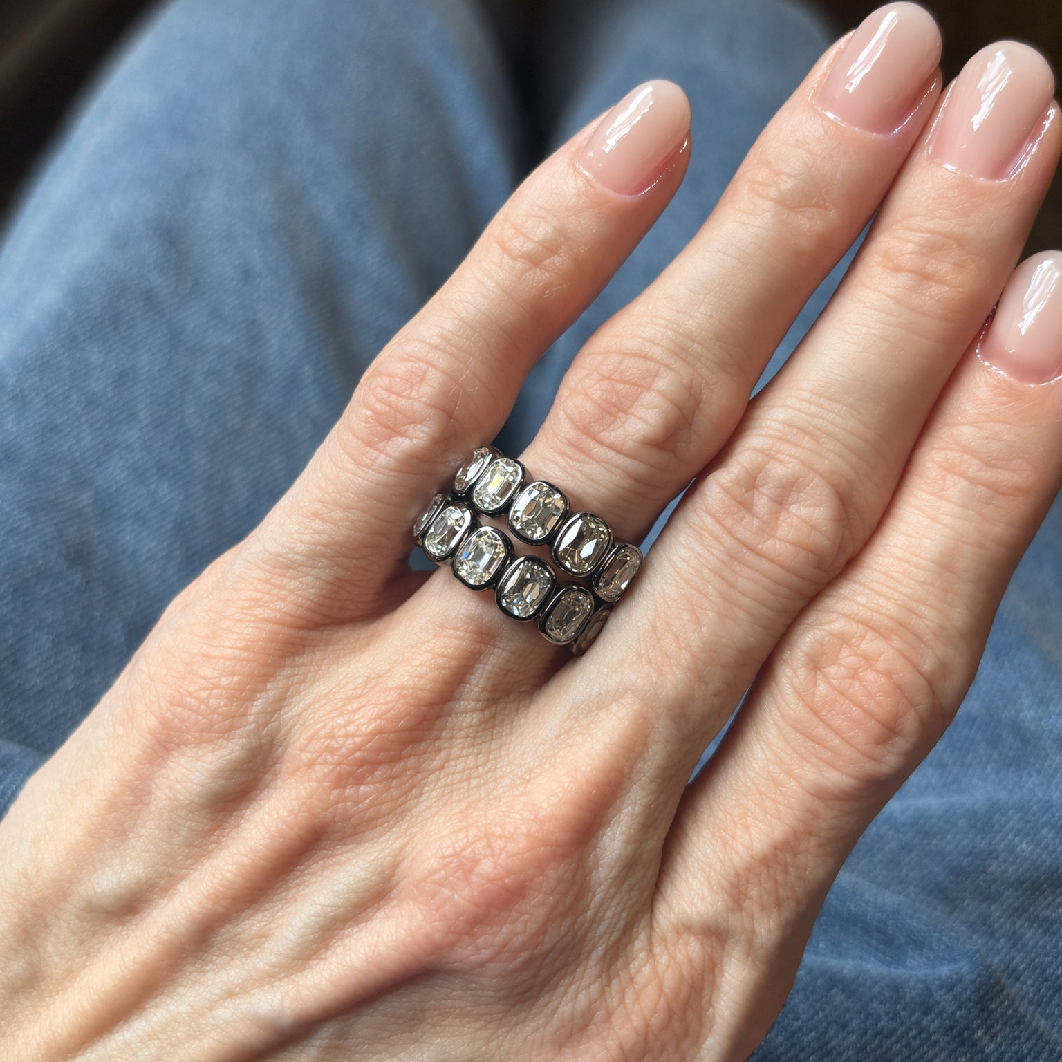 A hand with neatly manicured nails wears two silver rings with rectangular clear stones, complemented by the Bezel Set Cushion Cut Diamond Eternity Band in Black Rhodium, which features VS clarity stones. The person is wearing blue jeans in the background.