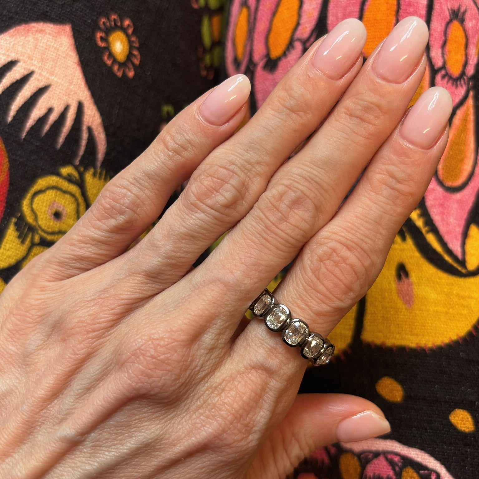 A hand with light pink manicured nails wears the Bezel Set Cushion Cut Diamond Eternity Band in Black Rhodium, featuring stunning cushion cut diamonds. The background displays colorful, floral-patterned fabric in shades of pink, yellow, and black.