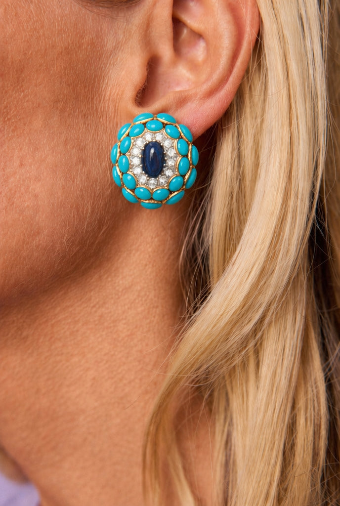 Close-up of a woman's ear adorned with a Van Cleef & Arpels pair of Sapphire, Turquoise and Diamond Earclips, featuring large round earrings set with turquoise stones around the edge, a central dark blue sapphire cabochon, and small white diamonds; her blonde hair is partially visible.