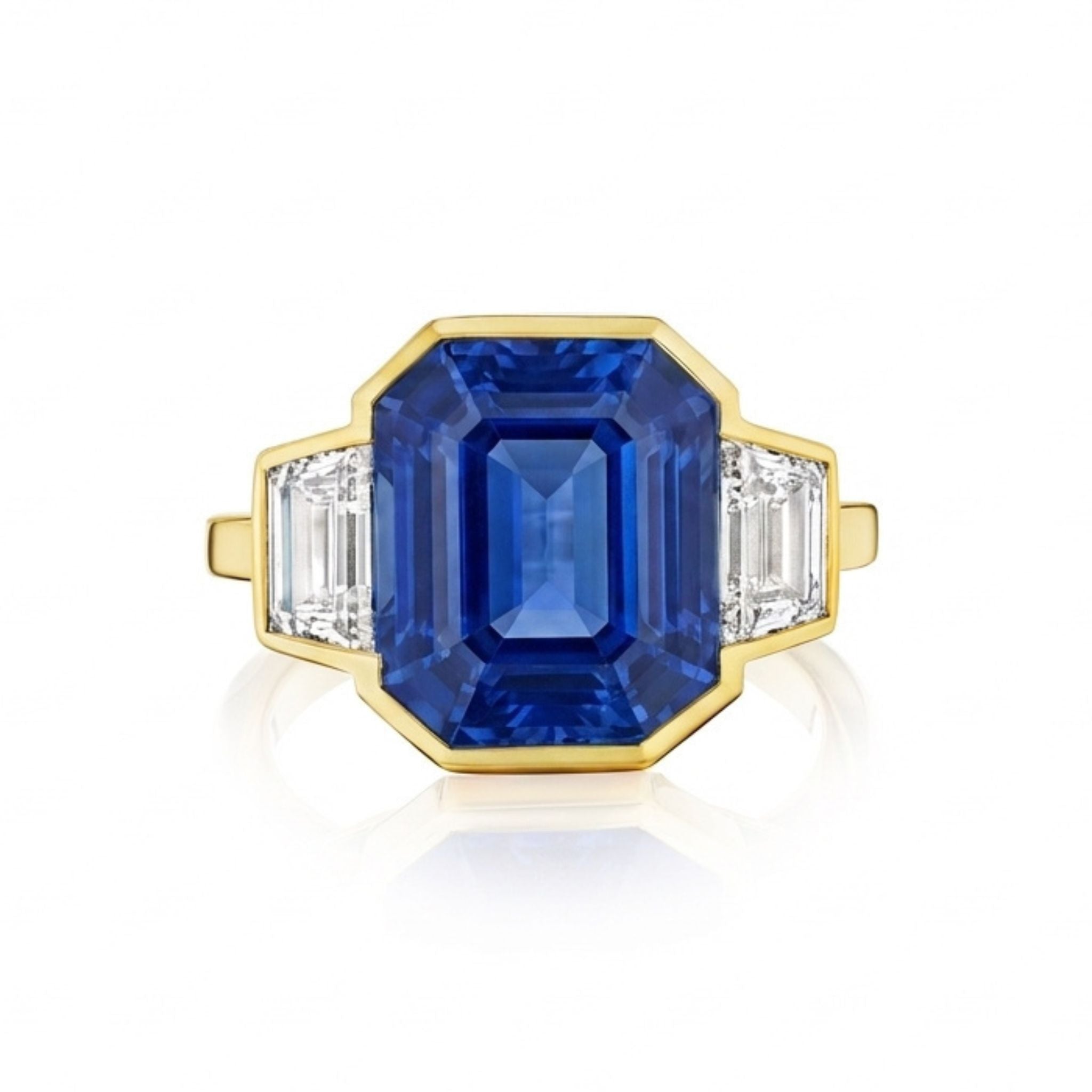 The Bezel Set Blue Sapphire Ring in yellow gold features an emerald-cut sapphire center and trapezoid diamond side stones, set against a white background.
