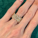 A hand with neatly manicured nails showcases the Multi Tier Marquise Diamond Eternity Band—an 18k gold ring adorned with multiple SI clarity marquise diamonds arranged in a leaf-inspired pattern—set against a green textured background.