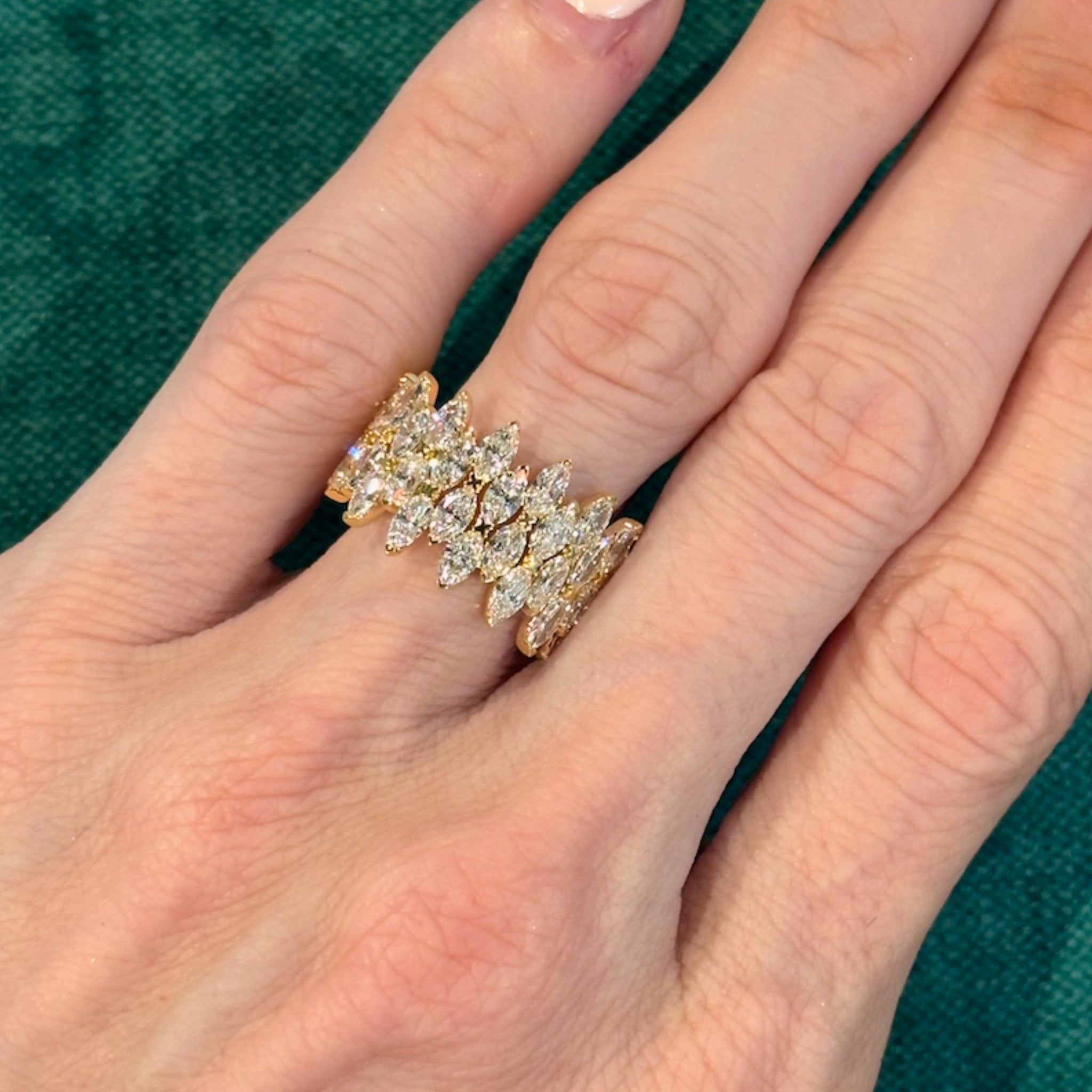A hand with neatly manicured nails showcases the Multi Tier Marquise Diamond Eternity Band—an 18k gold ring adorned with multiple SI clarity marquise diamonds arranged in a leaf-inspired pattern—set against a green textured background.