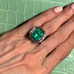 A hand with light pink nails wears the Mosaic Asscher Cut Emerald and Baguette Diamond Cocktail Ring in Platinum, featuring a striking emerald set in platinum and accented by shimmering baguette diamonds, resting on a light green, crocodile-textured surface.