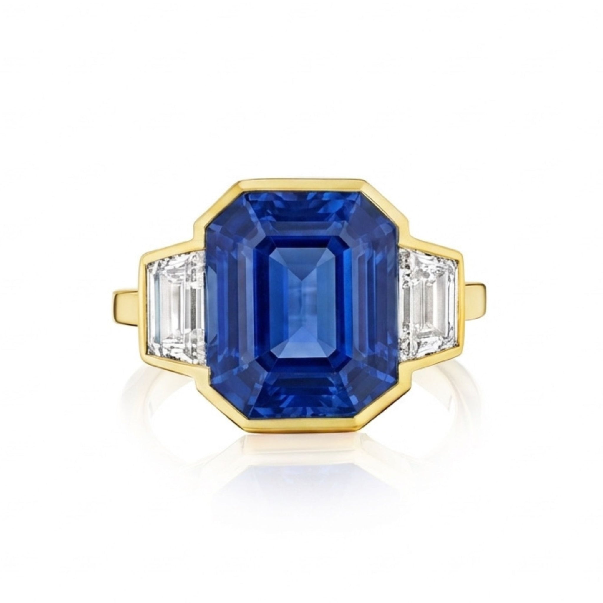 The Bezel Set Blue Sapphire Ring with Diamond Trapezoid Side Stones in Yellow Gold features a striking emerald-cut blue sapphire at its center, bezel set in 18k yellow gold and accented by two sparkling trapezoid diamond side stones.