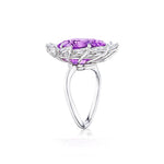 A side view of the Sunburst Cocktail Ring showcases its 18k white gold band, featuring a large oval purple sapphire centerpiece set in an intricate floral-inspired design and accented with a halo of small marquise diamonds, displayed against a white background.