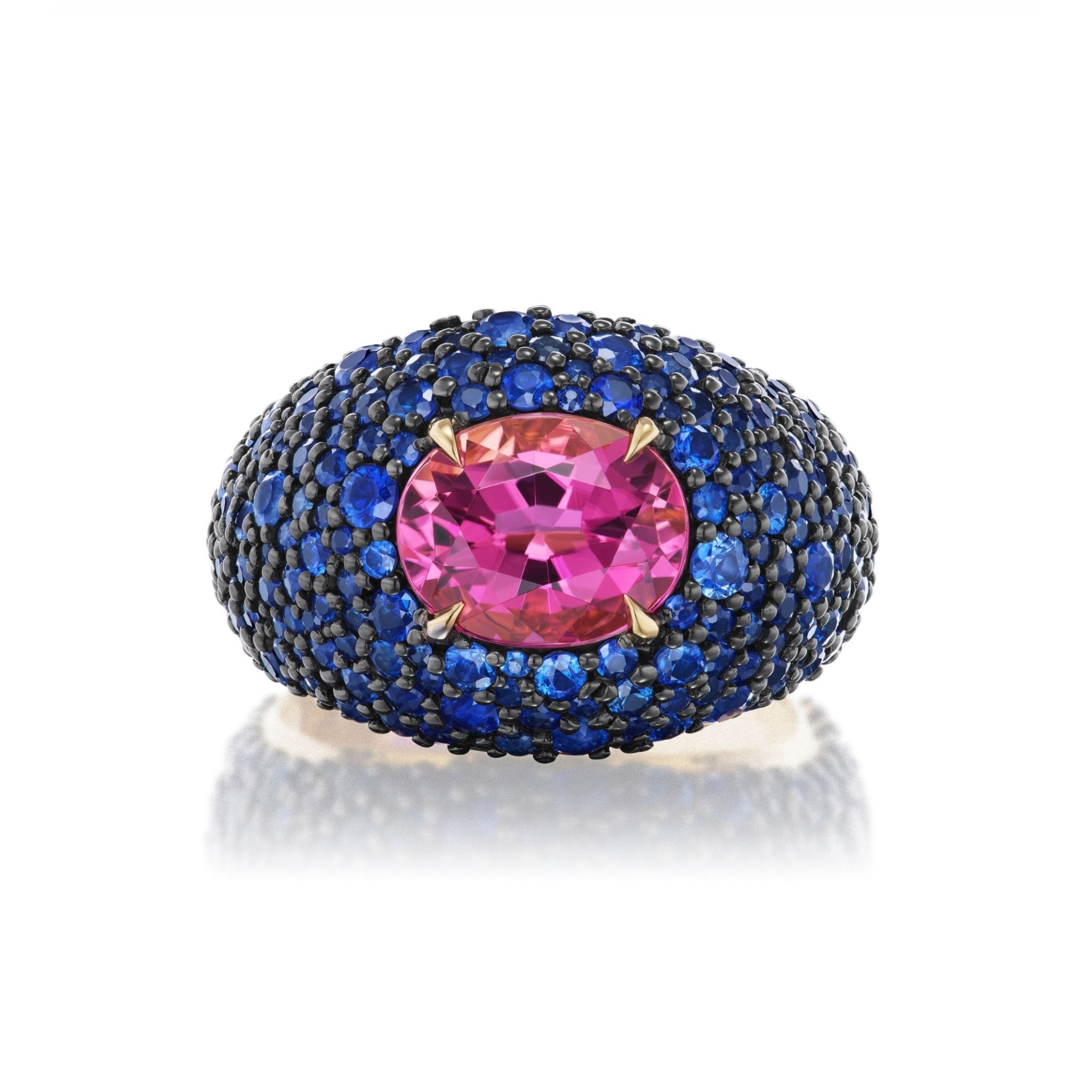 The Cobblestone Bombé Ring in Yellow Gold with Oval and Round Sapphires features a striking large oval hot pink sapphire at its center, surrounded by closely set small blue sapphires in a domed design. Displayed against a white background, the vibrant gemstones are beautifully complemented by an elegant 18k yellow gold setting.