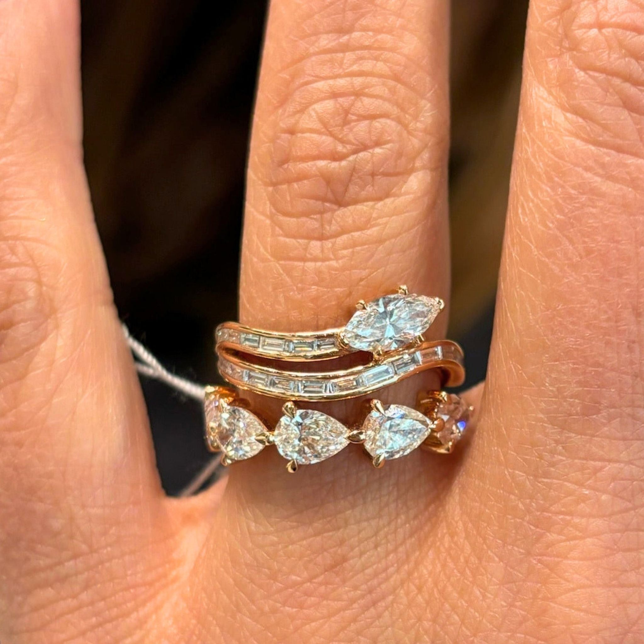 A hand wearing two 18k yellow gold rings: one, the Snake Wave Channel Set Baguette Diamond Ring with Marquise Diamond in Yellow Gold, is adorned with baguette and marquise-shaped diamonds; the other features pear-shaped diamonds—both stacked elegantly on a finger.