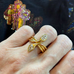A hand wearing the Double-Headed Serpent Ring with Pear Diamonds, crafted in 18k yellow gold with two pear-shaped diamonds set on overlapping gold bands. The background features black fabric adorned with a colorful, iridescent floral pattern.
