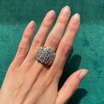 A hand with neatly manicured nails wears the Hexagon Bezel Set Multi-Tier Chunky Band in yellow gold, featuring multiple hexagonal shapes each bezel-set with sparkling round diamonds, against a green textured background.