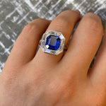 A hand is shown wearing the Mosaic Ring, which features an Asscher cut blue sapphire at the center surrounded by clear baguette diamonds, set in 18k gold, against a textured gray background.