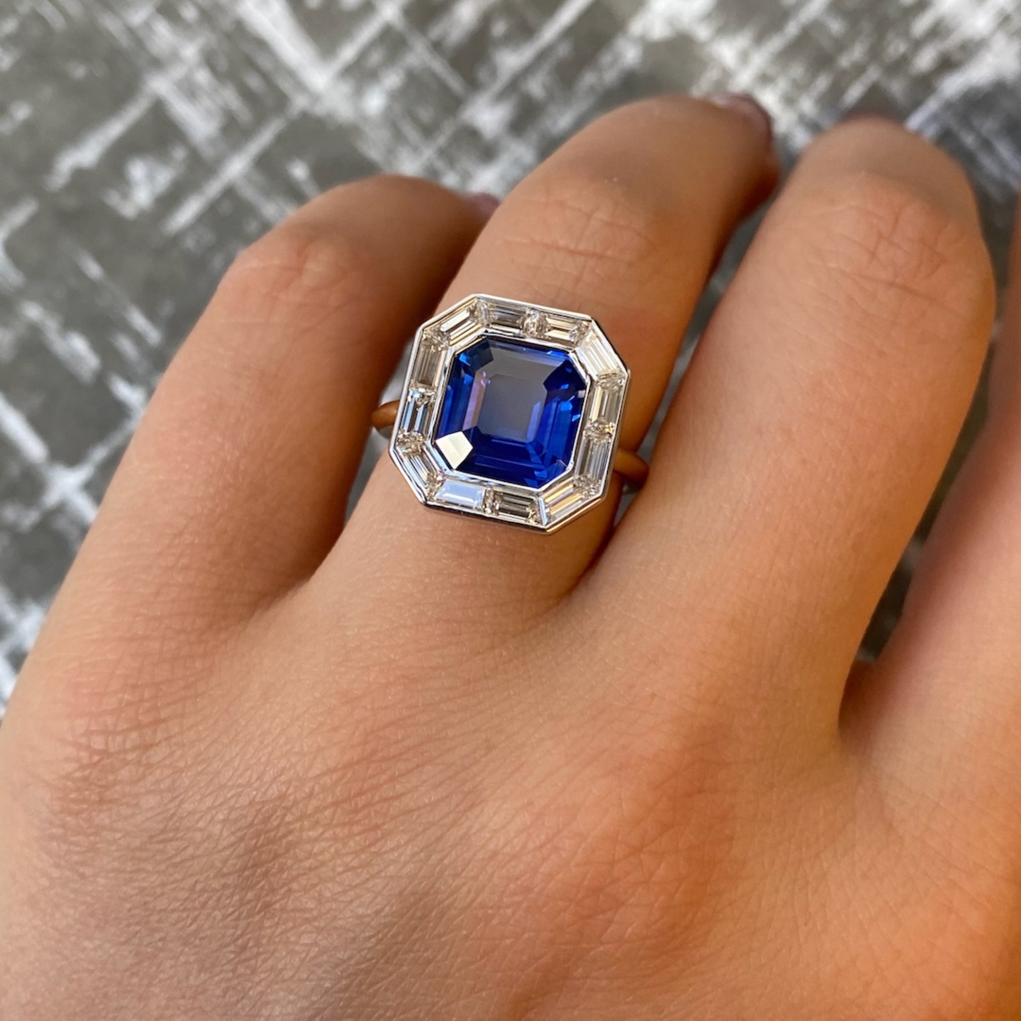 A hand is shown wearing the Mosaic Ring, which features an Asscher cut blue sapphire at the center surrounded by clear baguette diamonds, set in 18k gold, against a textured gray background.