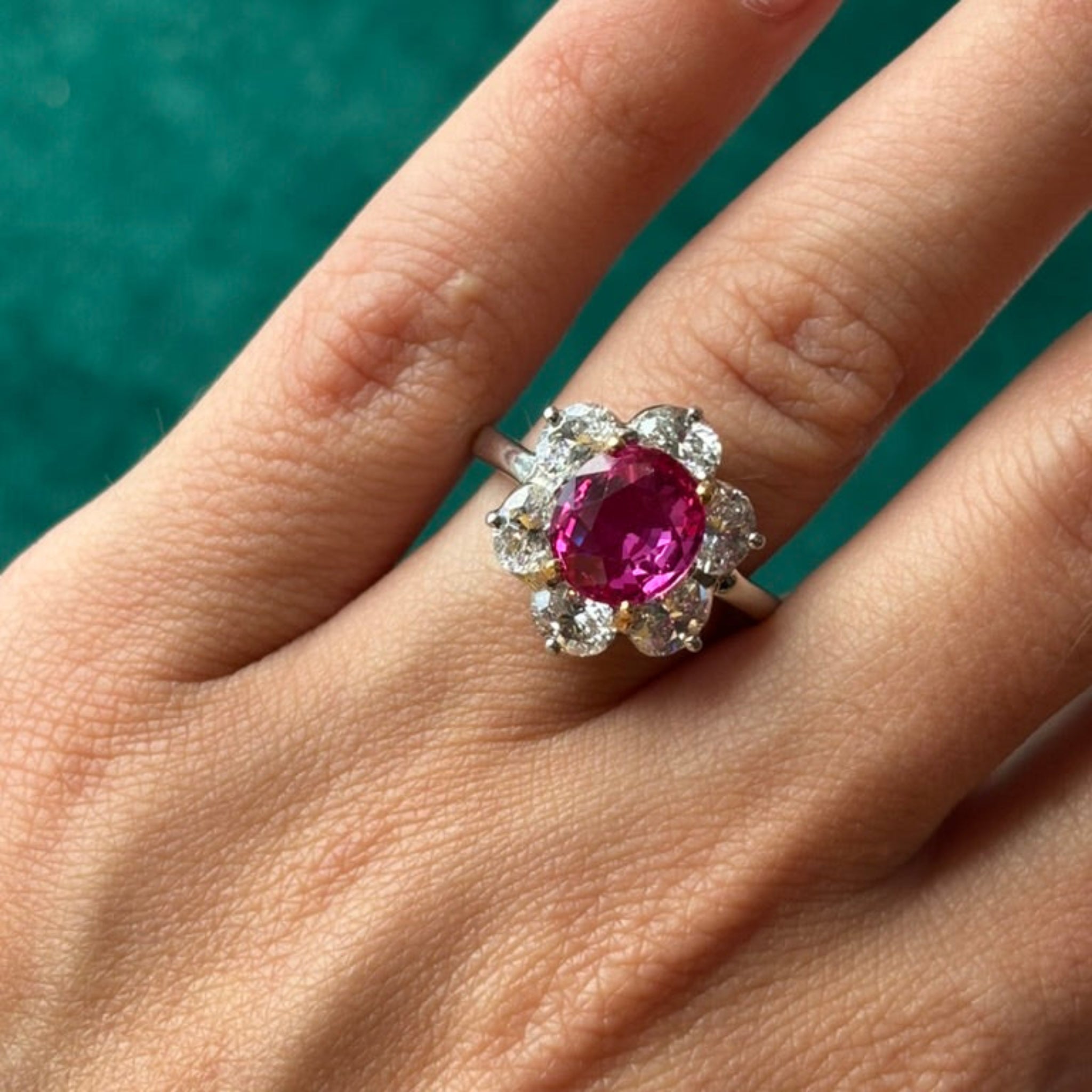 A hand displays the Oscar Heyman & Brothers Pink Sapphire and Diamond Ring, featuring a large oval Ceylon pink sapphire surrounded by clear diamonds, set in platinum white gold against a green background.
