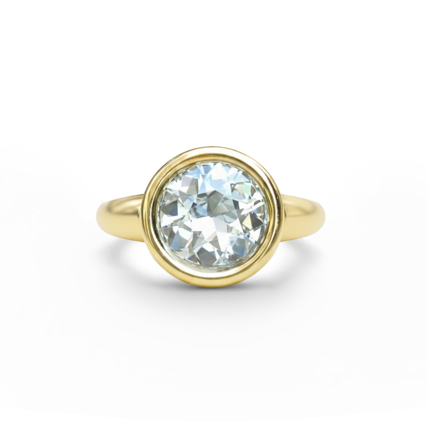 The Bezel Set Round Diamond Engagement Ring in Yellow Gold features a large, round diamond in a smooth bezel setting, shown against a white background.