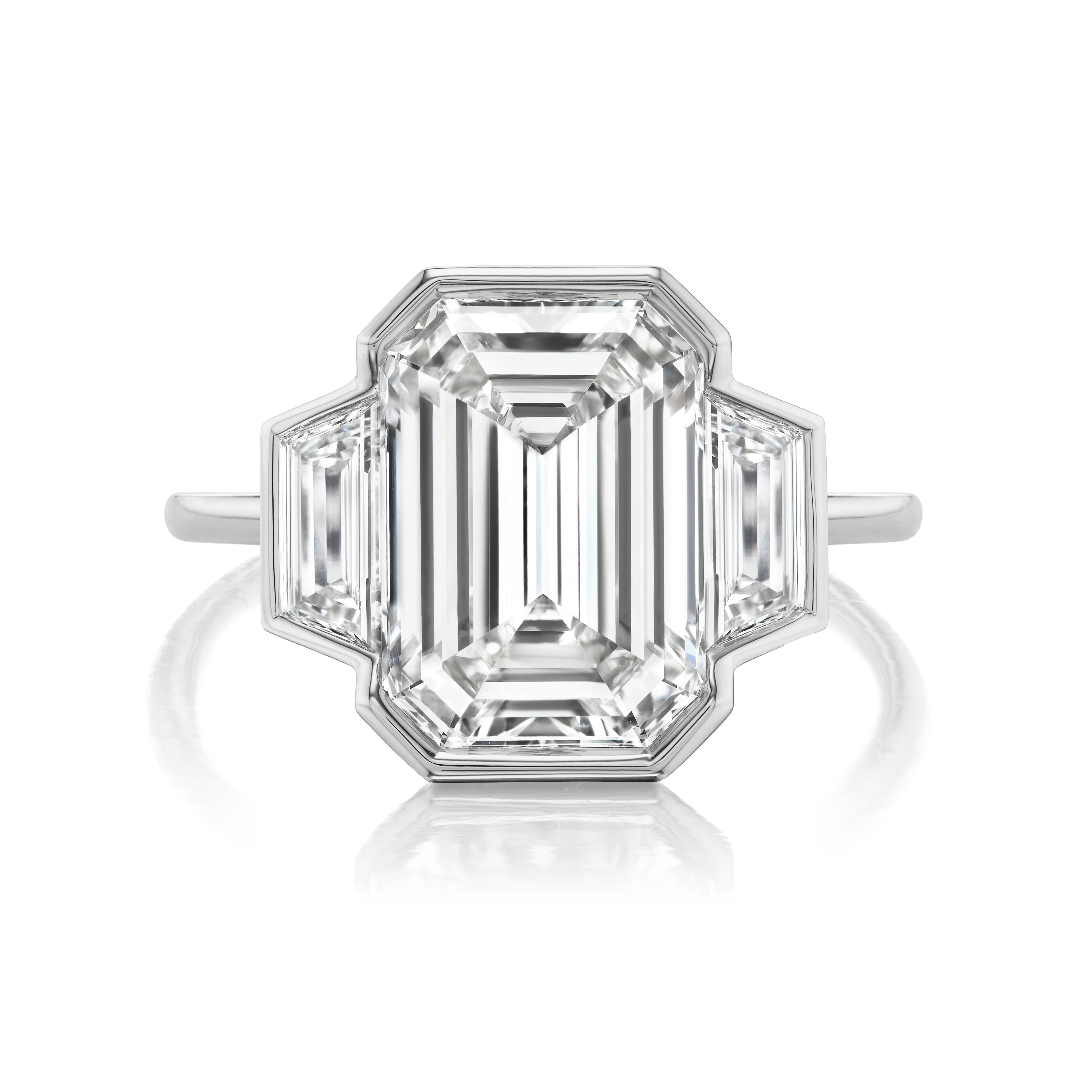 The Half Bezel Set Three Stone Engagement Ring features an emerald cut diamond center with trapezoid side stones in a geometric platinum setting and slim band.