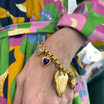 A hand in a colorful patterned dress wears the Gold, Colored Stone and Diamond Charm Bracelet, featuring puffy heart charms, including a blue enamel heart and an engraved gold locket.