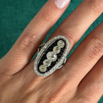 A hand with light skin wears the Onyx and Diamond Ring, an oval piece featuring five old mine-cut diamonds in a row, bordered by calibré-cut onyx and smaller diamonds, all crafted in platinum 1930s style.